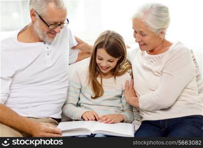 family, generation and people concept - smiling grandfather, granddaughter and grandmother with book sitting on couch at home