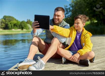 family, generation and technology concept - happy father and son with tablet pc computer sitting on river berth. happy father and son with tablet pc on river berth
