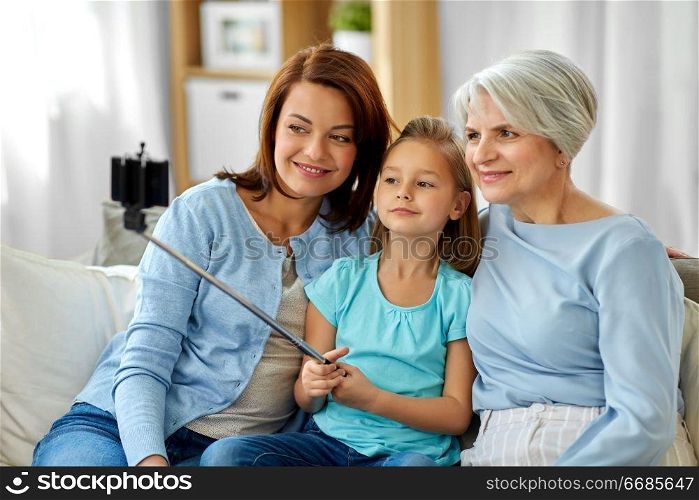 family, generation and technology concept - happy mother, daughter and grandmother taking picture by smartphone on selfie stick at home. mother, daughter and grandmother taking selfie