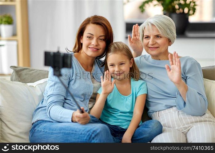 family, generation and technology concept - happy mother, daughter and grandmother taking picture by smartphone on selfie stick and waving hands at home. mother, daughter and grandmother taking selfie