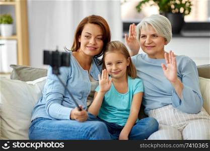 family, generation and technology concept - happy mother, daughter and grandmother taking picture by smartphone on selfie stick and waving hands at home. mother, daughter and grandmother taking selfie