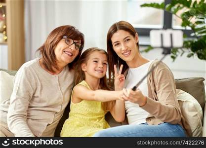 family, generation and technology concept - happy mother, daughter and grandmother taking picture by smartphone on selfie stick at home. mother, daughter and grandmother taking selfie