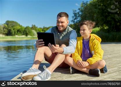 family, generation and technology concept - happy smiling father and son with tablet pc computer sitting on river berth. happy father and son with tablet pc on river berth