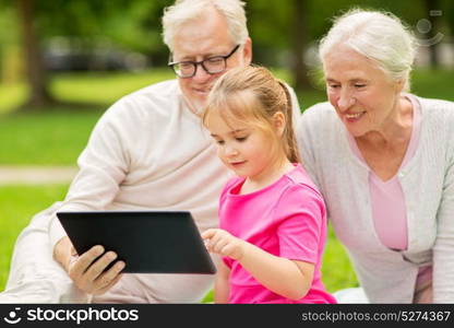 family, generation and technology concept - happy smiling grandmother, grandfather and little granddaughter with tablet pc computer sitting on blanket at park. grandparents and granddaughter with tablet pc