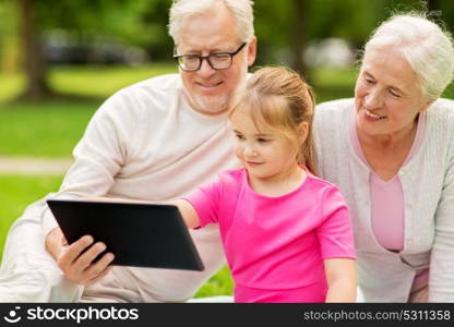 family, generation and technology concept - happy smiling grandmother, grandfather and little granddaughter with tablet pc computer sitting on blanket at park. grandparents and granddaughter with tablet pc