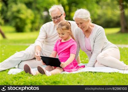 family, generation and technology concept - happy smiling grandmother, grandfather and little granddaughter with tablet pc computer sitting on blanket at park. grandparents and granddaughter with tablet pc