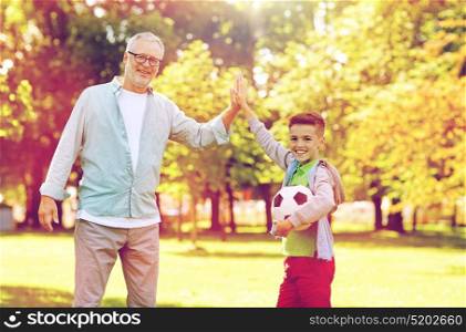 family, generation, gesture, sport and people concept - happy grandfather and grandson with football ball making high five at at summer park. old man and boy with soccer ball making high five