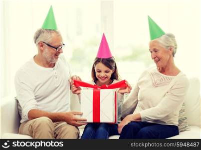 family, generation, holidays and people concept - smiling grandfather, granddaughter and grandmother in party hats with gift box at home