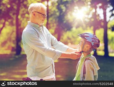 family, generation, safety and people concept - happy grandfather helping boy with bike helmet at summer park. old man helping boy with bike helmet at park