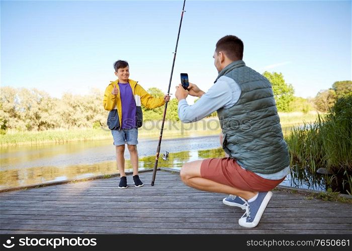 family, generation, summer holidays and people concept - father with smartphone photographing his happy smiling son with fishing rod showing thumbs up on river. father photographing son with fishing rod on river