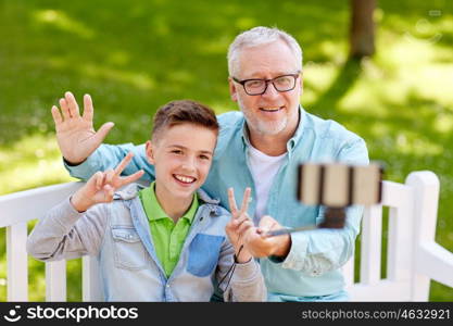 family, generation, technology and people concept - happy grandfather and grandson taking picture with smartphone selfie stick at summer park