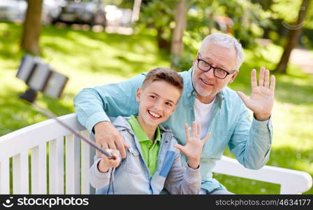 family, generation, technology and people concept - happy grandfather and grandson taking picture with smartphone selfie stick at summer park