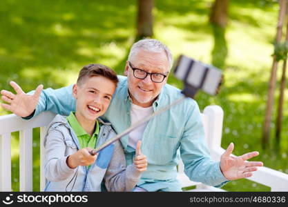 family, generation, technology and people concept - happy grandfather and grandson taking picture with smartphone selfie stick at summer park