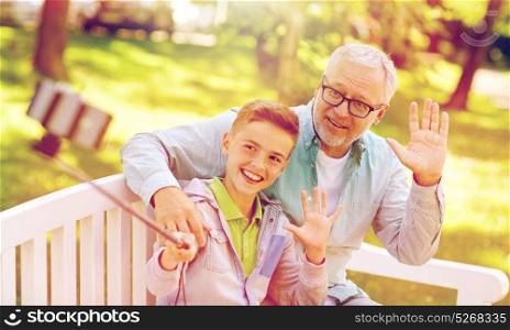 family, generation, technology and people concept - happy grandfather and grandson taking picture with smartphone selfie stick at summer park. old man and boy taking selfie by smartphone