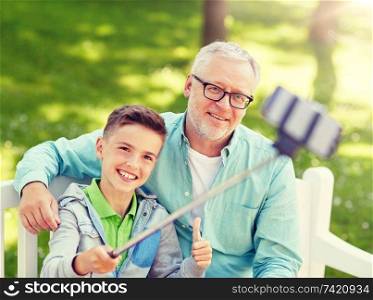 family, generation, technology and people concept - happy grandfather and grandson taking picture with smartphone selfie stick at summer park. old man and boy taking selfie by smartphone