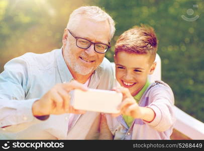 family, generation, technology and people concept - happy grandfather and grandson with smartphone taking selfie at summer park. old man and boy taking selfie by smartphone