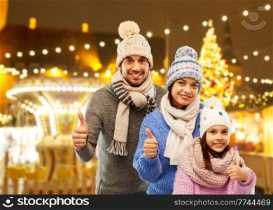 family, gesture and winter holidays concept - happy mother, father and little daughter in knitted hats and scarves showing thumbs up over night christmas market and lights background. happy family in winter clothes on grey background