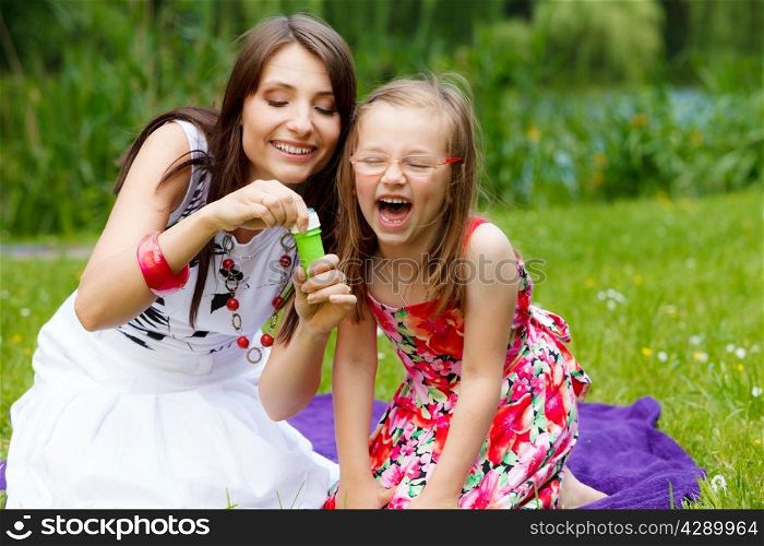 Family happiness and carefree concept. Mother and daughter little girl having fun blowing soap bubbles together in park, green blurred background