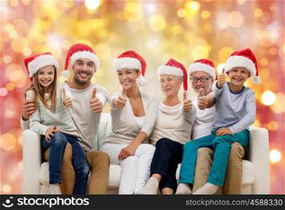 family, happiness, generation, holidays and people concept - happy family in santa helper hats sitting on couch and showing thumbs up gesture over lights background