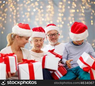 family, happiness, generation, holidays and people concept - happy family in santa helper hats with gift boxes sitting on couch at home over lights background