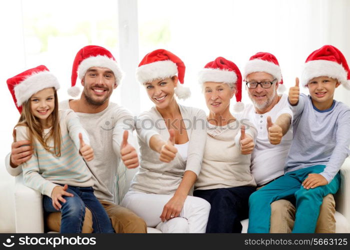 family, happiness, generation, holidays and people concept - happy family in santa helper hats sitting on couch and showing thumbs up gesture at home