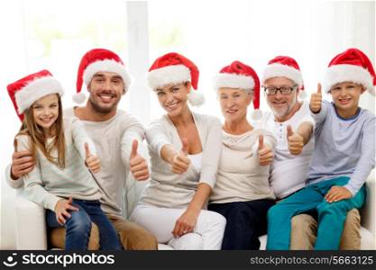 family, happiness, generation, holidays and people concept - happy family in santa helper hats sitting on couch and showing thumbs up gesture at home