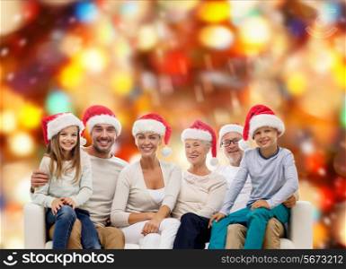 family, happiness, generation, holidays and people concept - happy family in santa helper hats sitting on couch over red lights background