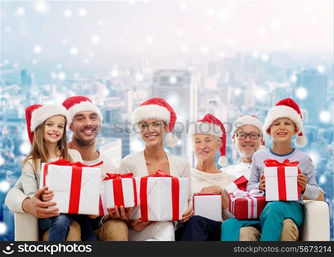 family, happiness, generation, holidays and people concept - happy family in santa helper hats with gift boxes sitting on couch over snowy city background