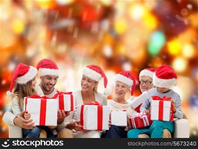 family, happiness, generation, holidays and people concept - happy family in santa helper hats with gift boxes sitting on couch over red lights background