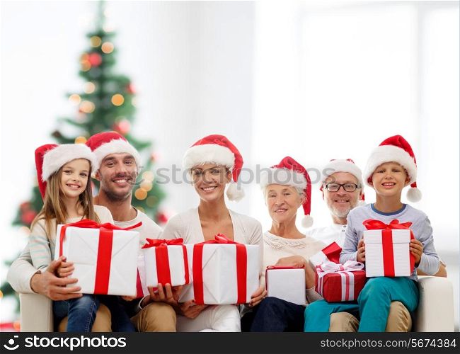family, happiness, generation, holidays and people concept - happy family in santa helper hats with gift boxes sitting on couch over living room and christmas tree background