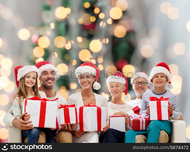 family, happiness, generation, holidays and people concept - happy family in santa helper hats with gift boxes sitting on couch over christmas tree lights background