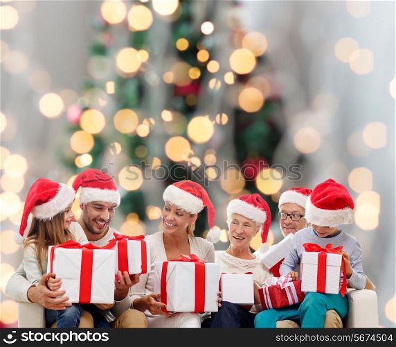 family, happiness, generation, holidays and people concept - happy family in santa helper hats with gift boxes sitting on couch over christmas tree lights background