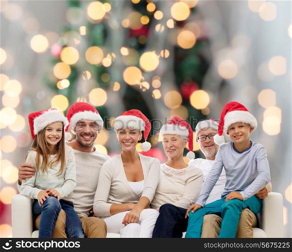 family, happiness, generation, holidays and people concept - happy family in santa helper hats sitting on couch over christmas tree lights background