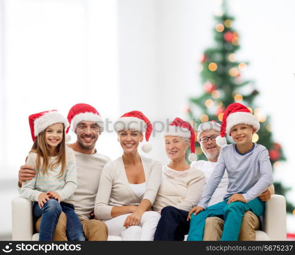 family, happiness, generation, holidays and people concept - happy family in santa helper hats sitting on couch over living room and christmas tree lights background