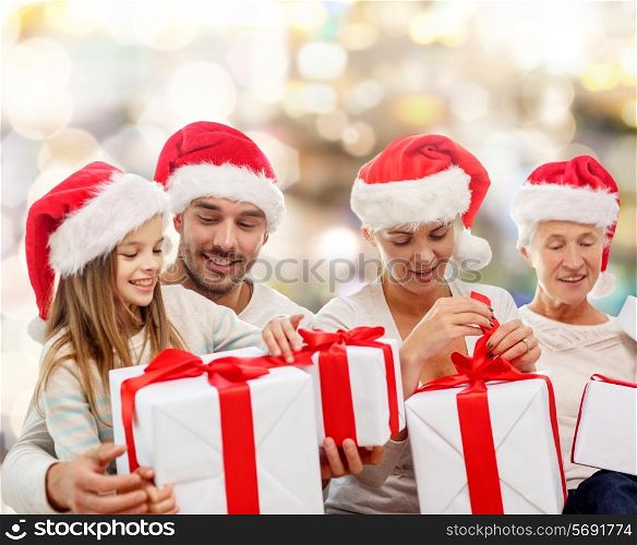 family, happiness, generation, holidays and people concept - happy family in santa helper hats with gift boxes sitting on couch over lights background