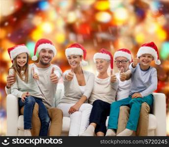 family, happiness, generation, holidays and people concept - happy family in santa helper hats sitting on couch and showing thumbs up gesture over red lights background