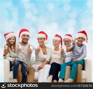 family, happiness, generation, holidays and people concept - happy family in santa helper hats sitting on couch and showing thumbs up gesture over blue lights background