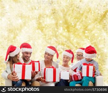 family, happiness, generation, holidays and people concept - happy family in santa helper hats with gift boxes sitting on couch over yellow lights background