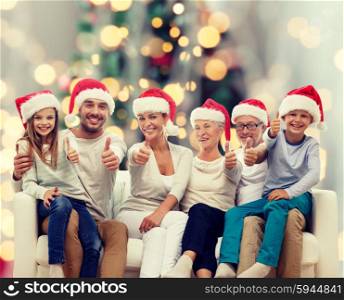 family, happiness, generation, holidays and people concept - happy family in santa helper hats sitting on couch and showing thumbs up gesture over tree lights background
