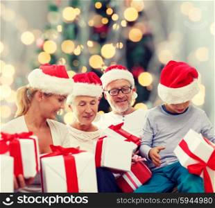 family, happiness, generation, holidays and people concept - happy family in santa helper hats with gift boxes sitting over christmas tree lights background