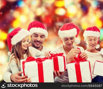 family, happiness, generation, holidays and people concept - happy family in santa helper hats with gift boxes sitting on couch over red lights background