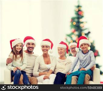 family, happiness, generation, holidays and people concept - happy family in santa helper hats sitting on couch over living room and christmas tree lights background