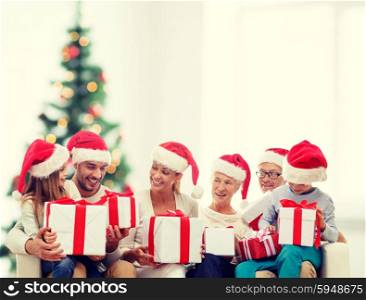 family, happiness, generation, holidays and people concept - happy family in santa helper hats with gift boxes sitting on couch over living room and christmas tree background