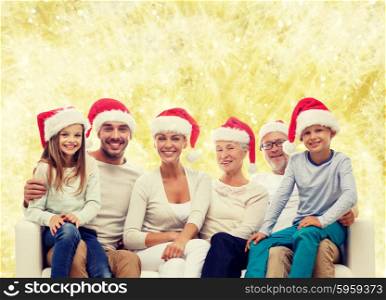 family, happiness, generation, holidays and people concept - happy family in santa helper hats sitting on couch over yellow lights background