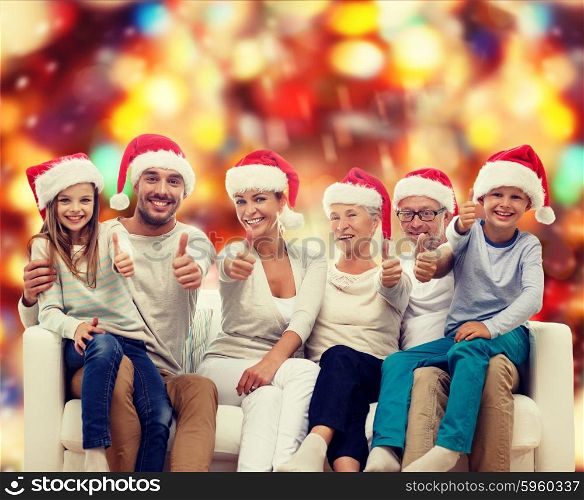 family, happiness, generation, holidays and people concept - happy family in santa helper hats sitting on couch and showing thumbs up gesture over red lights background