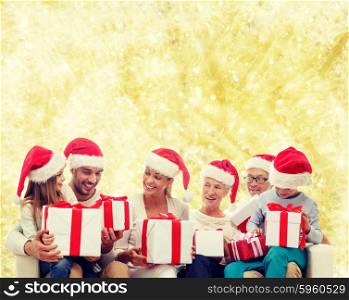 family, happiness, generation, holidays and people concept - happy family in santa helper hats with gift boxes sitting on couch over yellow lights background
