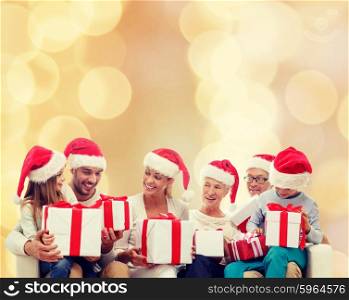 family, happiness, generation, holidays and people concept - happy family in santa helper hats with gift boxes sitting on couch over beige lights background