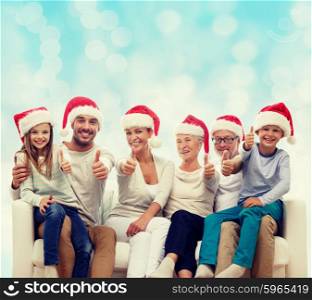family, happiness, generation, holidays and people concept - happy family in santa helper hats sitting on couch and showing thumbs up gesture over blue lights background