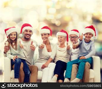 family, happiness, generation, holidays and people concept - happy family in santa helper hats sitting on couch and showing thumbs up gesture over lights background