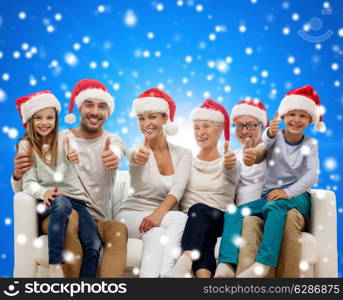 family, happiness, generation, holidays and people concept - happy family in santa helper hats sitting on couch and showing thumbs up gesture over blue snowy background